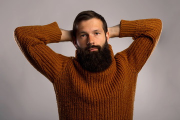 Handsome bearded man holds or scratch his nape, in casual warm knitted sweater putting both hands in his back of the head on gray background copy space