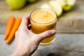 Wall Mural - Holding a glass of homemade freshly squeezed apple and carrot juice, apple and carrot on a side on the wooden rustic background