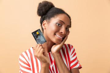 Joyous african american woman smiling and holding credit card, isolated over beige background