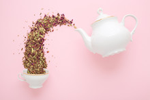 Dry Herbal Tea Pouring From White Porcelain Teapot Into Cup On Pink Surface. Flat Lay. Tea Time Concept.