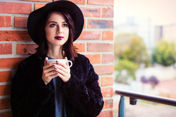 Wall Mural - Portrait of a young caucasian woman in black hat and coat with cup in autumn time on red brick wall background