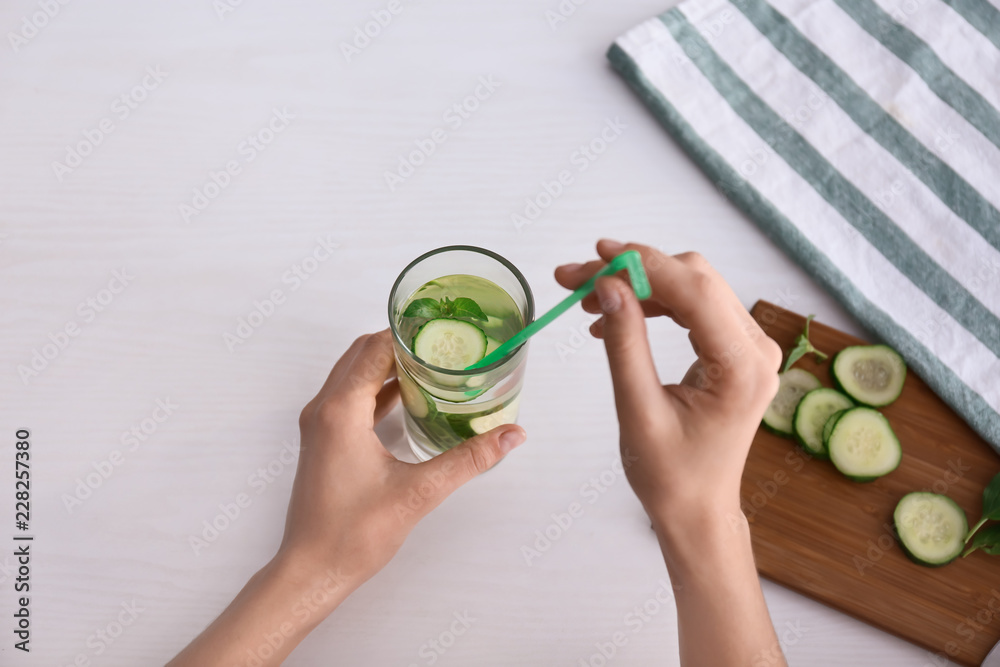 Woman putting straw into glass with cucumber water on light table