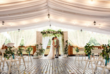 Wedding Ceremony. Elegant Wedding Couple Kissing Near Wedding Arch, Bride And Groom In Love