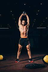 Wall Mural - handsome muscular man holding kettlebell overhead while working out  in dark gym