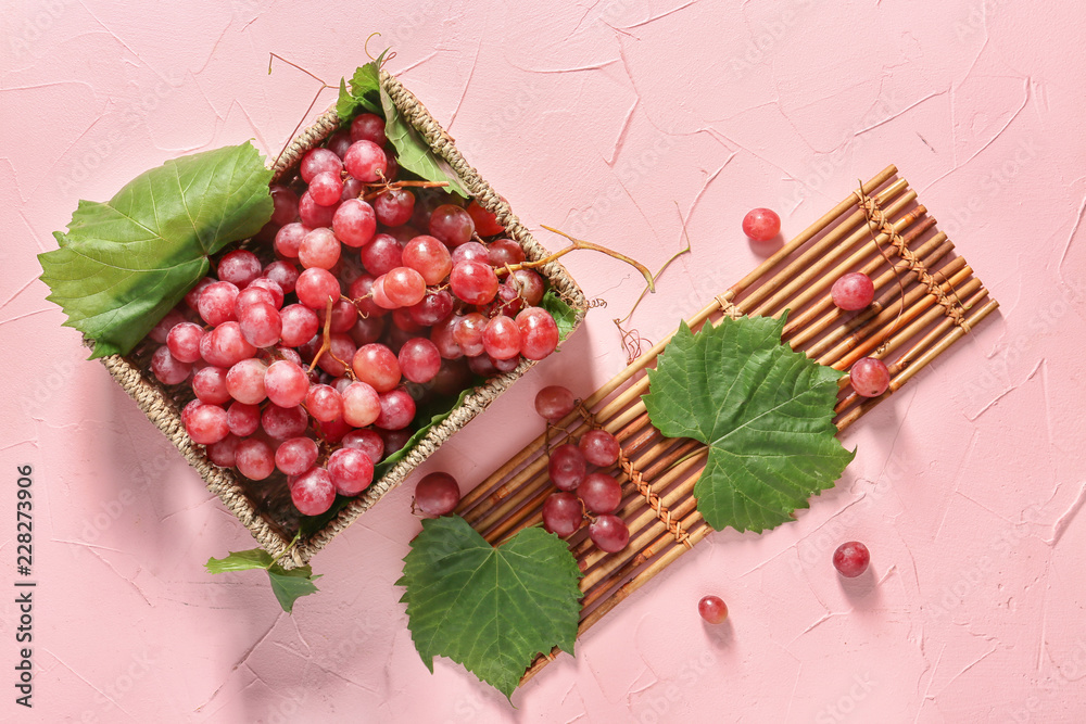 Wicker box with ripe grapes and bamboo mat on color background