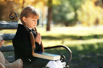 Little girl with Bible praying on bench outdoors