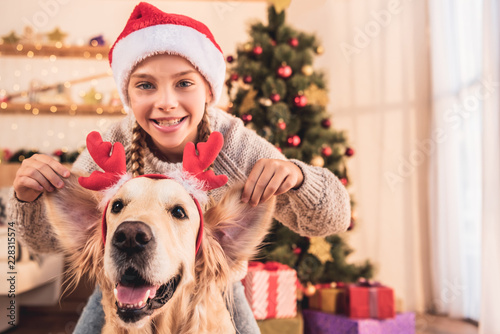 Fotografia smiling kid in santa hat and golden retriever dog with deer horns having fun at
