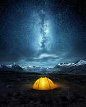 Camping In The Wilderness. A Pitched Tent Under The Glowing  Night Sky Stars Of The Milky Way With Snowy Mountains In The Background. Nature Landscape Photo Composite.