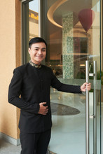 Asian Man In Elegant Black Suit Opening Glass Doorway Of Hotel Greeting Guests 