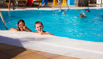 Little boy and girl with pink goggles smiling with thumbs up by swimming pool edge. Young kids having fun on summer holidays. Resort leisure time, travel vacation, hotel activity, tourism concepts