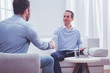 Glad to be helpful. Cheerful professional psychiatrist shaking hands with his satisfied client while sitting on the sofa and holding a clipboard in hand