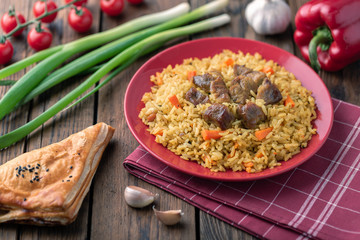 Wall Mural - Red plate with pilaf on a brown wooden table. On the table is red pepper, green onions, garlic, cherry tomatoes, red napkin, spoon.