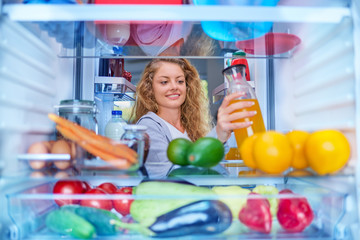 Wall Mural - Woman standing in front of fridge full of groceries and taking juice. Picture taken from inside of fridge.