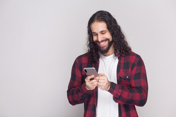 Cheerful handsome young man in red checkered shirt and long curly hair standing, using smartphone and texting message with toothy smile. Indoor, studio shot, isolated on grey background.