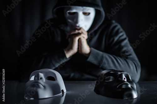 Hoodie man wearing mystery mask choosing black or white mask on the table. Anonymous social masking. Major depressive disorder or bipolar disorder. Halloween concept