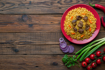 Wall Mural - Red plate with pilaf on a brown wooden table. On the table is red pepper, green onions, garlic, cherry tomatoes, red napkin, spoon. Top view. Flat lay.