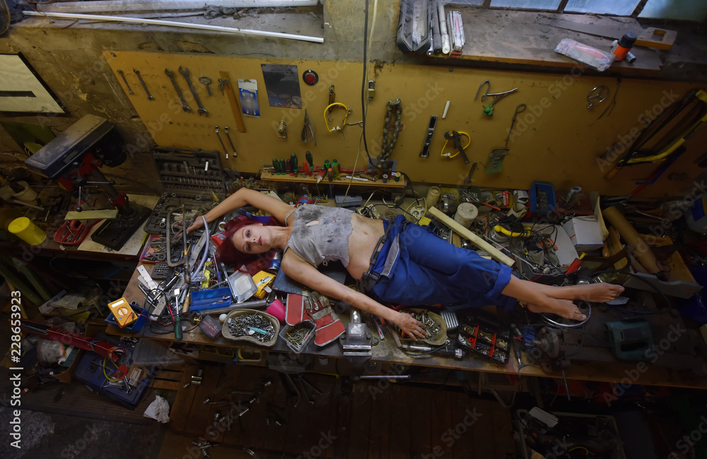 A young beautiful woman finds time to relax during  her working hours. She lies down on the work bench filled with tools and other electronic devices. - obrazy, fototapety, plakaty 