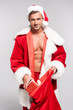 © LIGHTFIELD STUDIOS - sexy man in santa costume opening red bag and smiling at camera isolated on grey