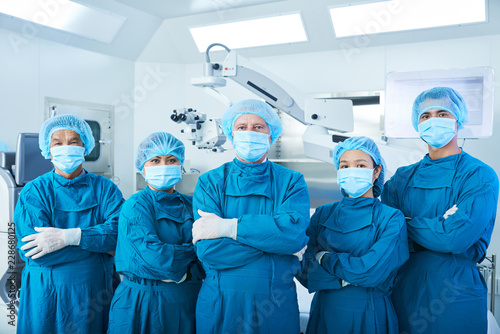 Fotografía Asian surgical team in masks and uniform keeping arms crossed and looking at cam