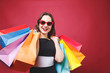 © proimagecontent - Young cute girl with bright makeup in black dress holding up many colorful paper shopping bags and looking at camera with smiling face on red background, black friday concept
