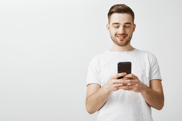Checking profile of girl he loves. Handsome delighted and happy caucasian guy with cool haircut and bristle holding smartphone looking at device screen with satisfaction reacting to daring message