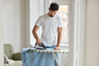 © WHstudio Leushin N - Photo of busy unshaven man irons shirt on ironing board, prepares for formal meeting on business conference, dressed in casual white t shirt, does her domestic duties, stands in spacious room