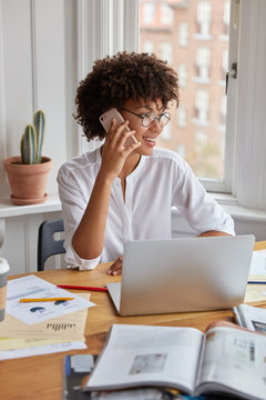 Vertical shot of pretty black businesswoman examines charts on papers, discusses future trading with business partner, works on portable laptop computer at workspace, looks positively aside.