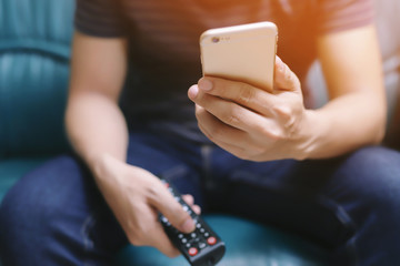 young man using television remote control and mobile smart phone is sitting on a sofa. on or off tv.