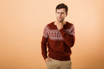 Canvas Print - Image of surprised man 20s with bristle wearing knitted sweater looking aside and touching chin, isolated over beige background