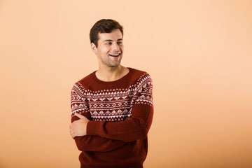 Image of young man 20s with bristle wearing knitted sweater posing on camera with hands crossed, isolated over beige background