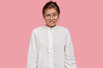 Wall Mural - Doubtful nervous woman bites lips with embarrassed expression, raises eyebrows, has to make serious decision, wears casual white shirt, spectacles, models against pink background. Facial expressions