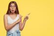 Studio shot of thrilled young lovely female model points witth both index fingers at upper right corner, discusses amazed advertisement, drops jaw with amazement, isolated over yellow studio wall