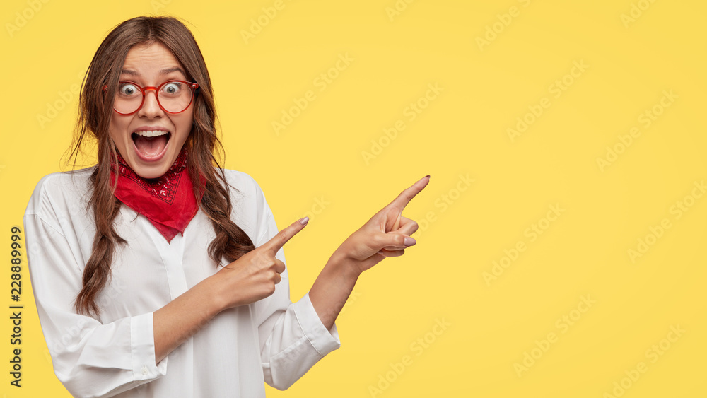 Amazed attractive student indicates with both index fingers aside, dressed in white shirt, poses over yellow background, gazes at camera, keeps mouth opened, wears fashionable clothes. Wow, look there - obrazy, fototapety, plakaty 