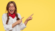Amazed Attractive Student Indicates With Both Index Fingers Aside, Dressed In White Shirt, Poses Over Yellow Background, Gazes At Camera, Keeps Mouth Opened, Wears Fashionable Clothes. Wow, Look There