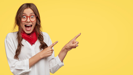 Amazed attractive student indicates with both index fingers aside, dressed in white shirt, poses over yellow background, gazes at camera, keeps mouth opened, wears fashionable clothes. Wow, look there