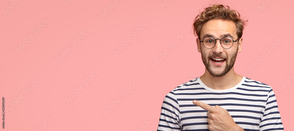 Cheerful curly man with bristle, points left, wears casual clothes and spectacles, has satisfied expression, shows free space for your slogan, promotional content or advertisemet. Look aside there - obrazy, fototapety, plakaty 