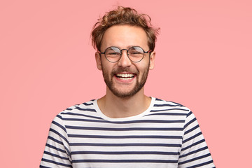 headshot of pleased hipster has satisfied expression, curly hair and bristle, wears round transparen