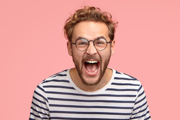Poster - Crazy man yells loudly, has overjoyed facial expression, shouts for his favourite team, wears round transparent glasses and striped clothes, poses over pink background. Amazed hipster exclaims