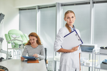 Wall Mural - Waist up portrait of beautiful lady in white lab coat standing and holding patient medical history. Red-haired woman sitting at the table and looking at documents on background