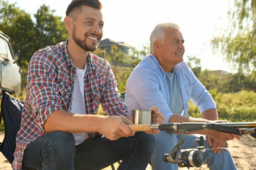 Father and adult son fishing together from riverside on sunny day