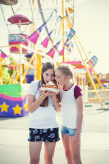 Two teen girls making a silly face while eating a funnel cake at an amusement park ride. Sticking out their tongues at pulling a funny face. Carefree and fun-loving teen girls at a carnival