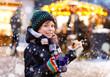 Little cute kid boy eating German sausage and drinking hot children punch on Christmas market. Happy child on traditional family market in Germany, Munich. Laughing boy in colorful winter clothes