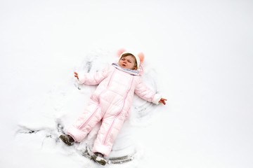 Wall Mural - Cute little baby girl in colorful winter clothes making snow angel, laying down on snow. Active outdoors leisure with children in winter. Toddler in pink snowsuit.