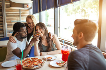 Cheerful happy male and female students treating each other with organic pizza with different toppings and non alchoholic cocktails, having fun and enjoying meal and carefree talks.