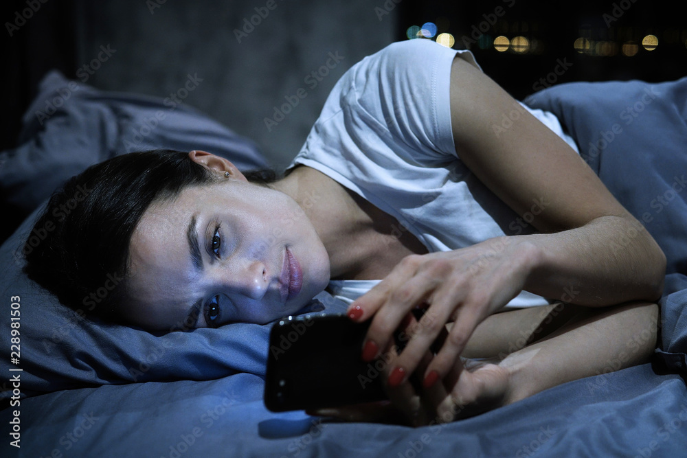 Smartphone addiction. Young tired female looking at her mobile phone screen, lying in bed late at night, scrolling her social media news feed