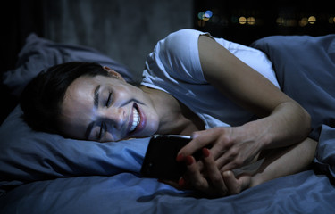 Dating app concept. Young woman lying in bed at night, looking at smartphone, laughing, texting