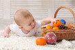 Infant boy on white wrap dressed in white touches mandarin in basket, christmas ball foreground