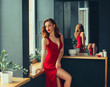 hot young adult, proud and domineering woman dressed in a long scarlet red long dress, sexually demonstrates her naked graceful leg, crouching on the window sill, room in loft stile with mirrors
