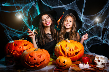 Happy Halloween . Beautiful caucasian mother and her daughter in witch costumes enjoy using magic with magic wand to halloween pumpkin jar over spider web on black studio background.