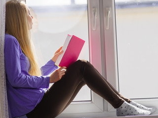 Woman sitting on window sill reading book at home